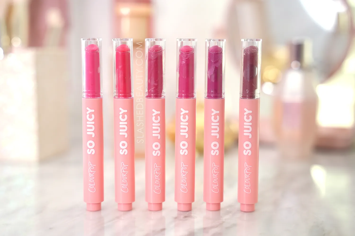 colourpop-so-juicy-gloss-balm-review | Slashed Beauty Six tubes of ColourPop So Juicy Plumping Gloss Balms standing side by side on a marble vanity | ColourPop So Juicy Gloss Balms Review | Slashed Beauty